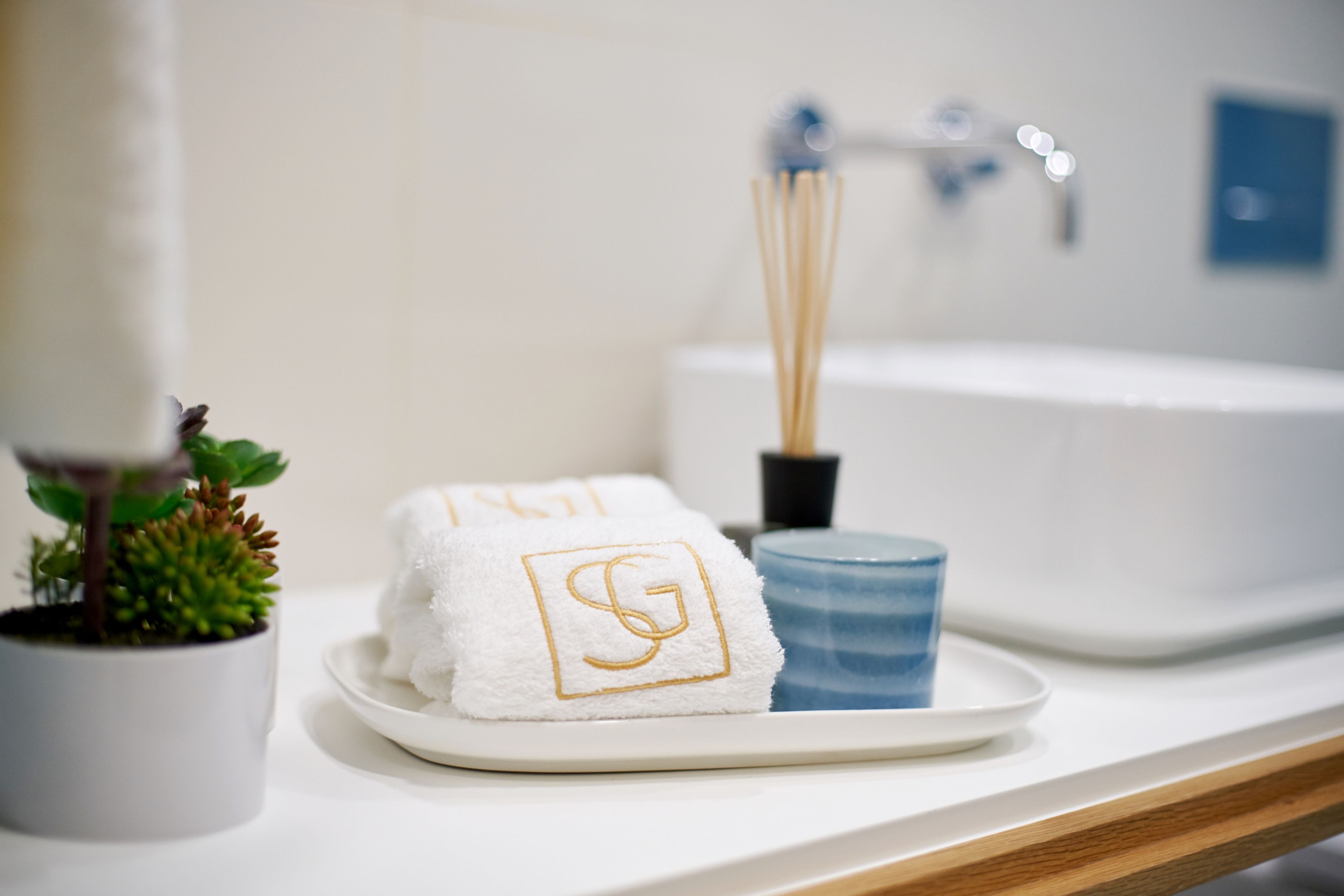 A bathroom with a white sink, a blue soap dish, a potted plant, and two towels with a monogram.
