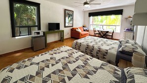 Ein Zimmer mit zwei Betten, einem Schreibtisch mit Computer, einem Fernseher, einem Deckenventilator und großen Fenstern mit Blick auf Grünflächen.