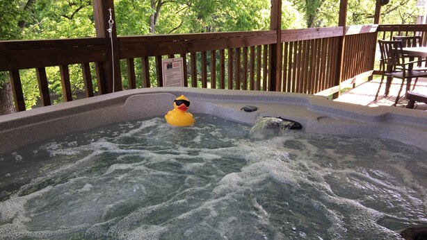 Un bain à remous avec un canard en plastique portant des lunettes de soleil, entouré d’une terrasse en bois et d’arbres.