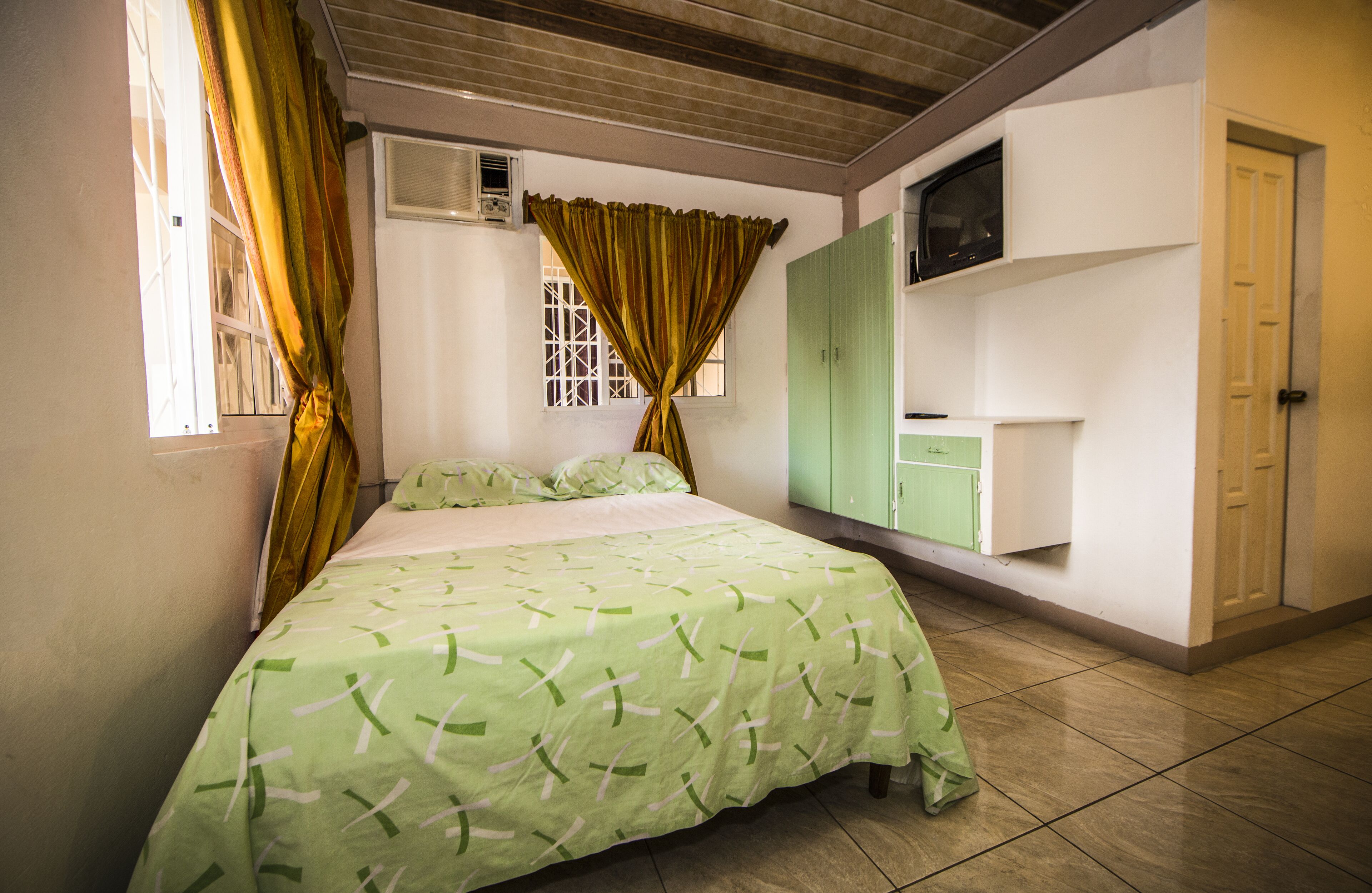 Una camera da letto con un letto, una finestra con tende, un condizionatore d'aria, un armadietto verde e una porta.