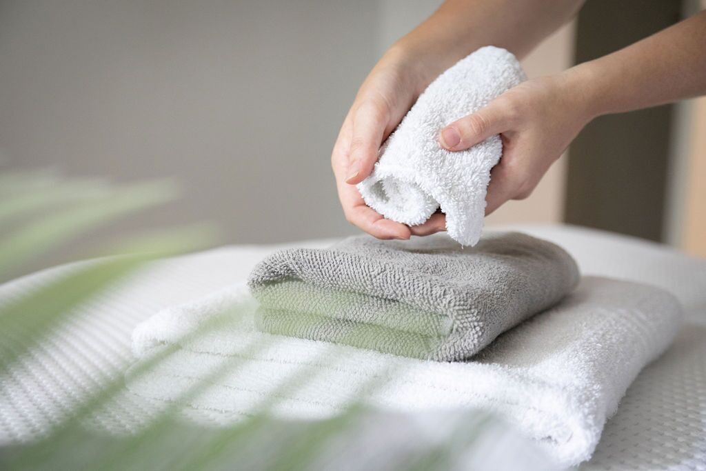 A hand holding a white towel over a stack of folded towels on a bed.