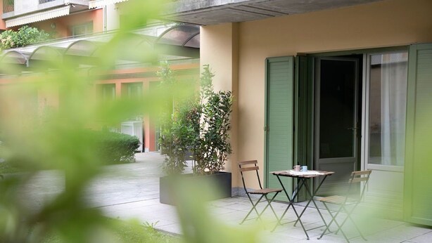 A balcony with a table and chairs, a potted plant, and a building with green shutters. - Donizetti Residence (Bergamo)