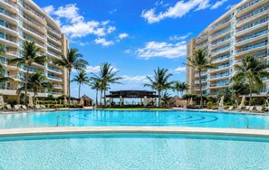 A resort pool with palm trees, a clear sky, and a building with multiple balconies. - Luxury Beachfront Condo at Del Canto Residences with Beautiful Ocean Views (Nuevo Vallarta)