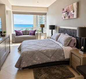 A bedroom with a large bed, a sofa, a nightstand, a lamp, and a view of the ocean.