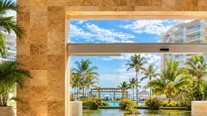 A modern hotel lobby with a large glass door leading to a pool area surrounded by palm trees and a clear sky. - Luxury Beachfront Condo at Del Canto Residences with Beautiful Ocean Views (Nuevo Vallarta)