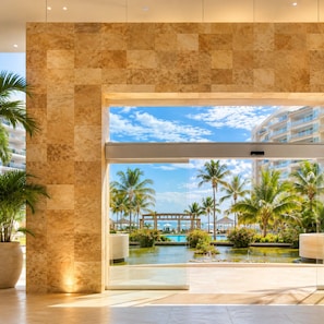A modern hotel lobby with a large glass door leading to a pool area surrounded by palm trees and a clear sky. - Luxury Beachfront Condo at Del Canto Residences with Beautiful Ocean Views (Nuevo Vallarta)