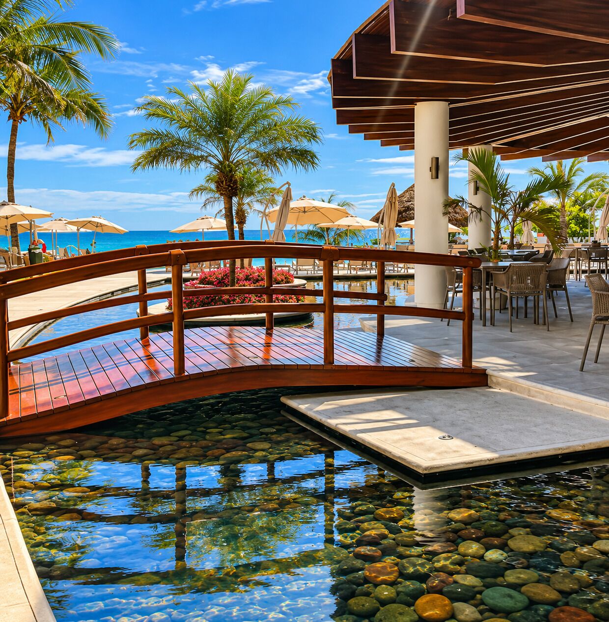 A wooden bridge over a pool with clear water, surrounded by palm trees and outdoor seating.