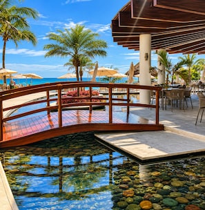 A wooden bridge over a pool with clear water, surrounded by palm trees and outdoor seating.