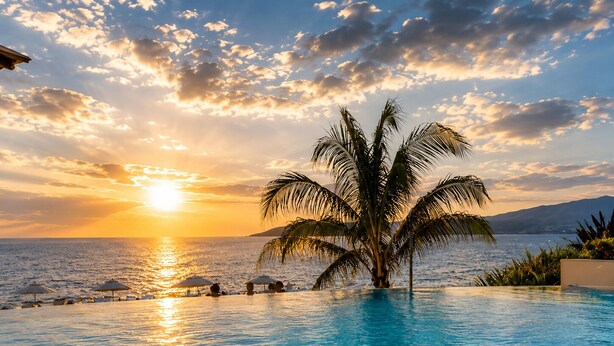 A pool with clear water reflecting the sunset, a palm tree, and a view of the sea and mountains. - Luxury Beachfront Condo at Del Canto Residences with Beautiful Ocean Views (Nuevo Vallarta)