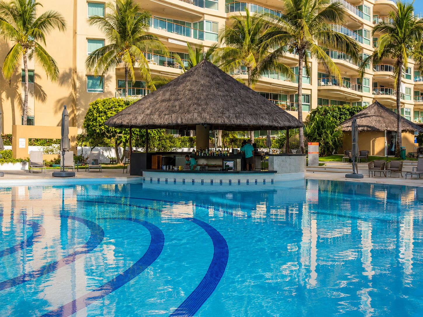 A swimming pool with a bar area, palm trees, and a multi-story hotel building in the background.