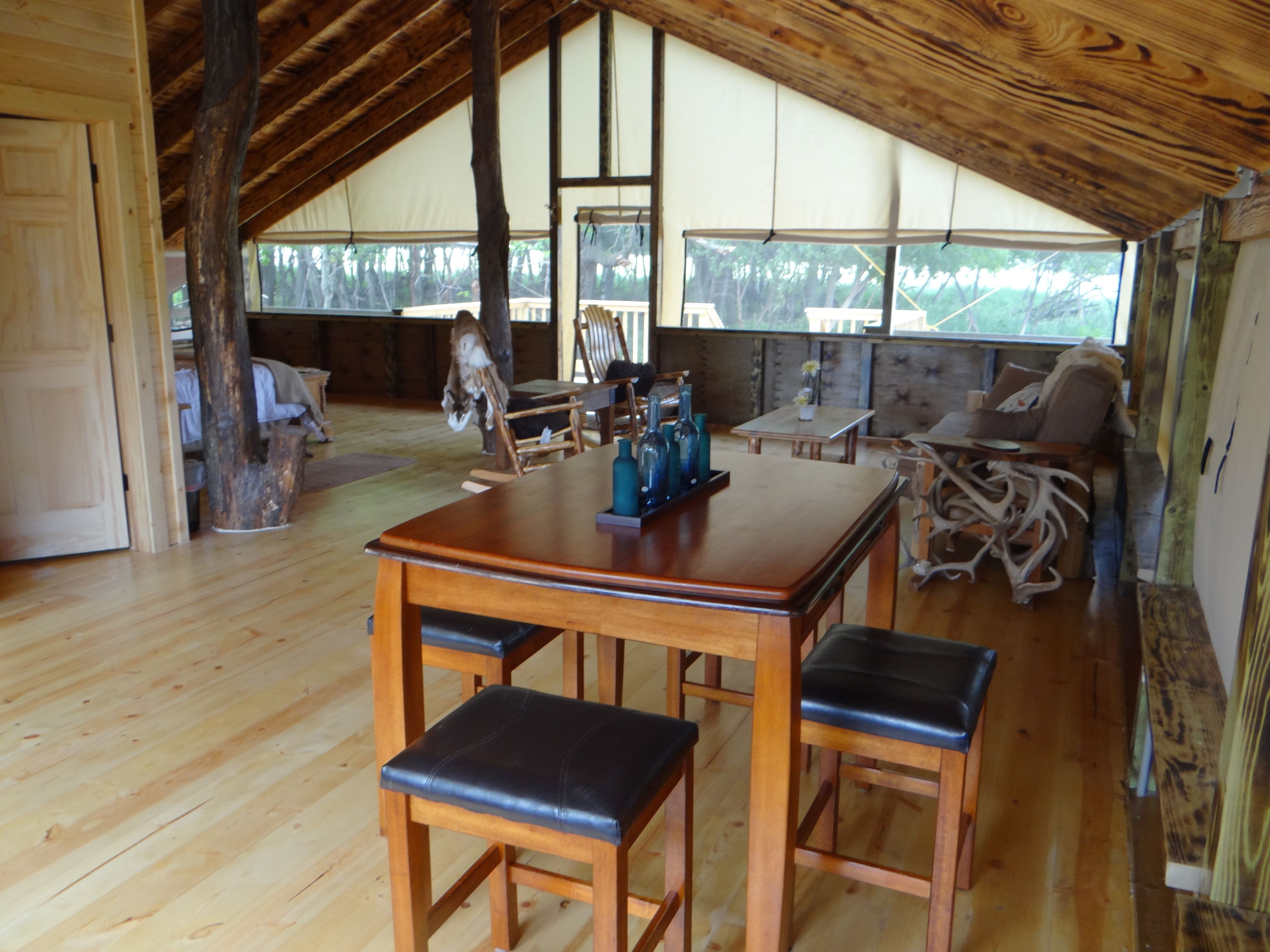 A rustic wooden interior with a high ceiling, a dining table with benches, and a view of the outdoors through large windows.