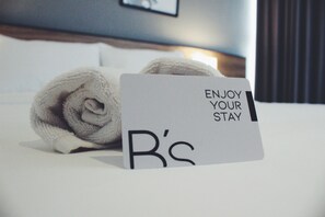 Una toalla doblada con cuidado y una tarjeta en una cama con el texto "ENJOY YOUR STAY" y "B's".