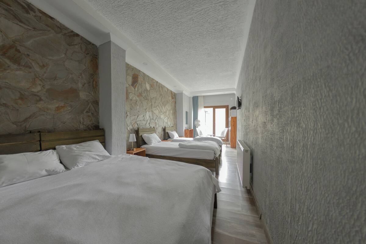 A hotel room with a stone wall, two beds, a wooden headboard, a nightstand, and a window with curtains.