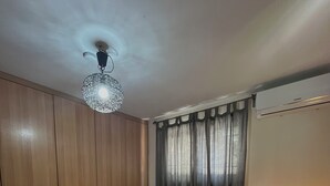 Quarto com cama, mesa de cabeceira com abajur, guarda-roupa, luminária no teto e janela com cortinas.