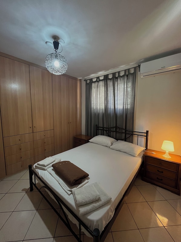 A bedroom with a bed, a bedside table with a lamp, a wardrobe, a ceiling light fixture, and a window with curtains.