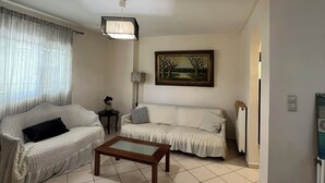 Uma sala de estar com dois sofás brancos, uma mesa de centro de madeira, um quadro na parede, um abajur e um radiador.