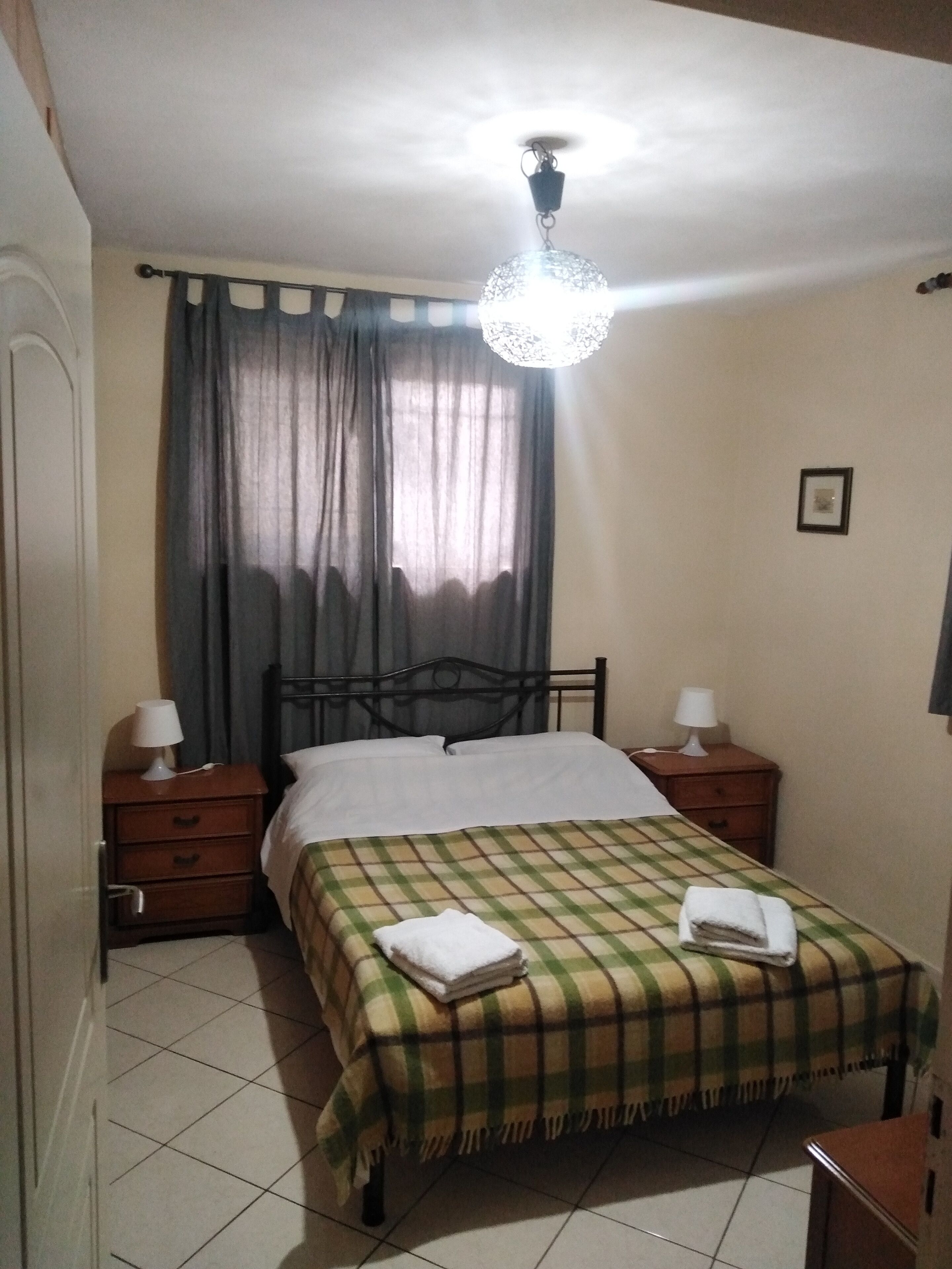 A bedroom with a bed, two bedside tables with lamps, a window with curtains, and a ceiling light fixture.