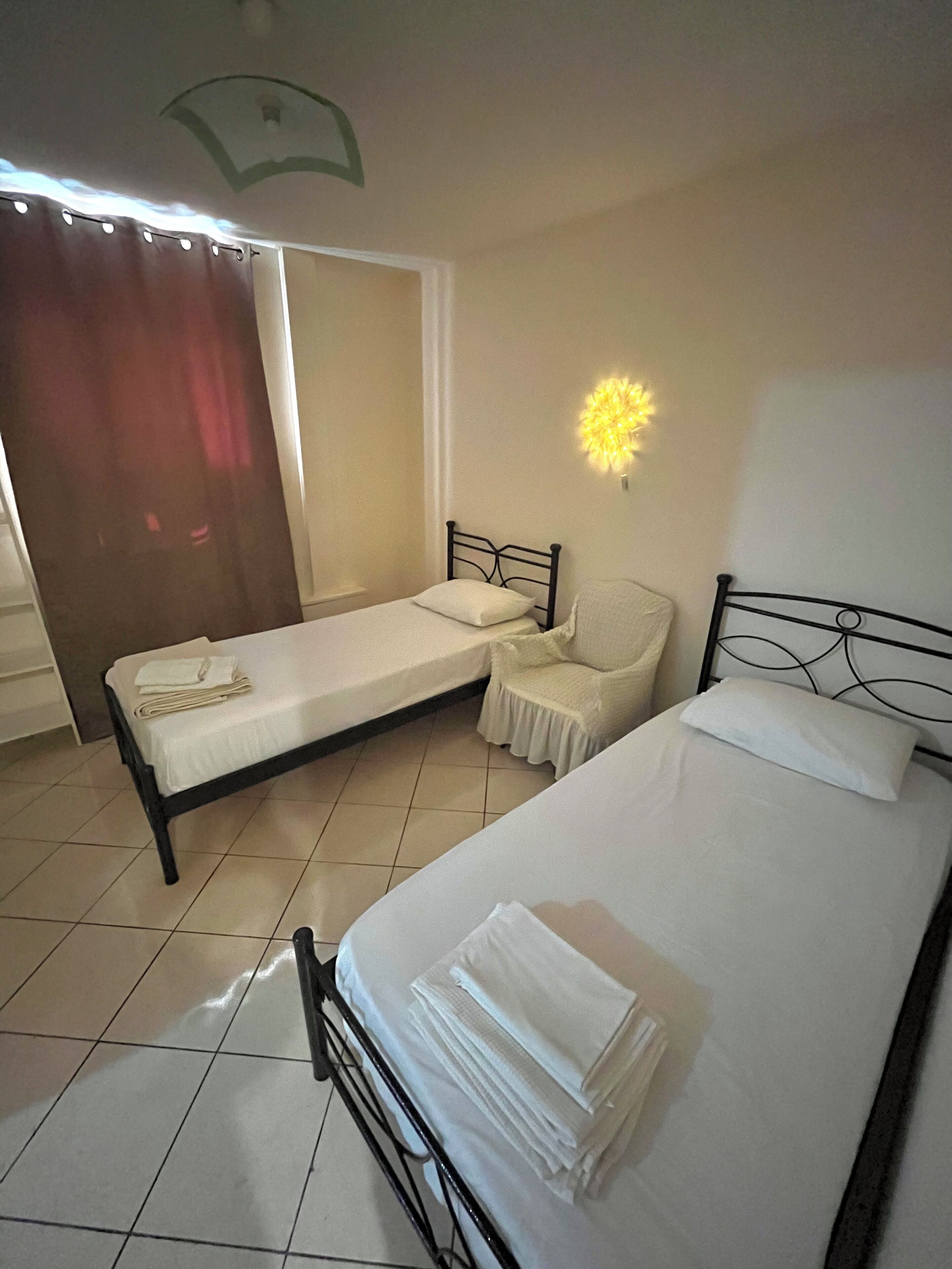 Una habitación con dos camas, una silla, una ventana con cortinas y una lámpara fijada a la pared.