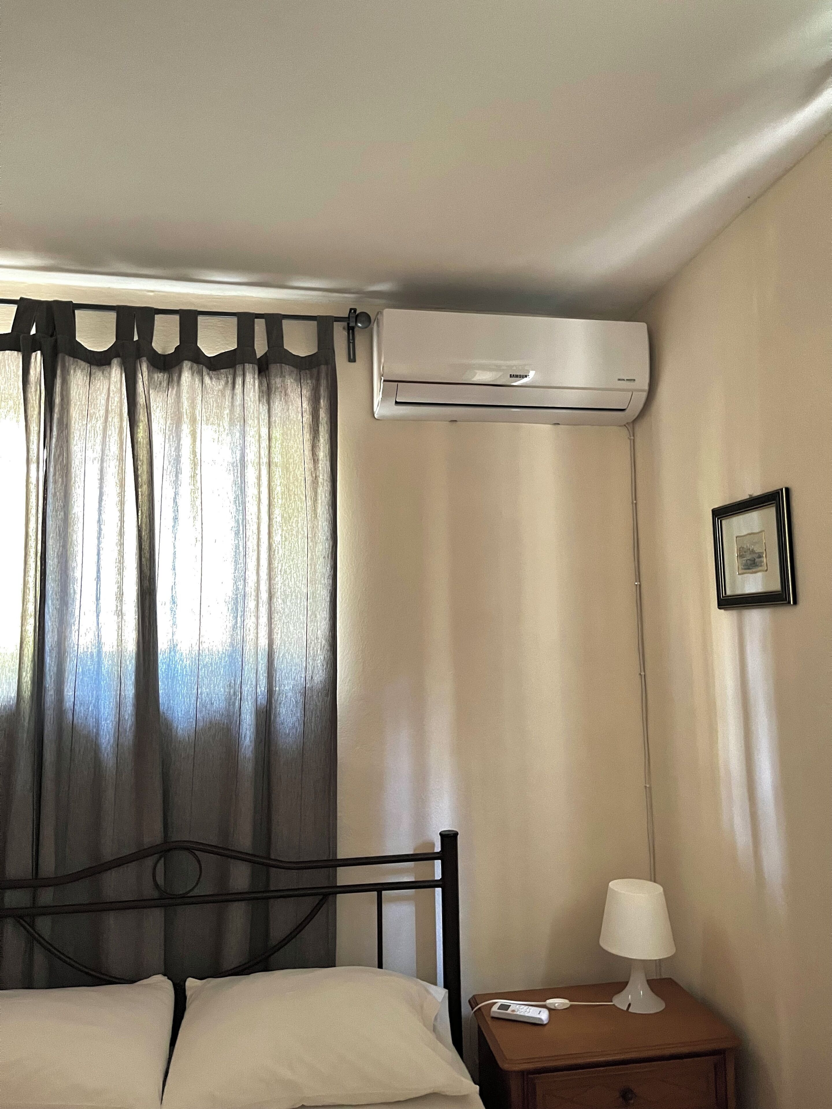 Una habitación de hotel con cama, unidad de aire acondicionado, mesita de noche con lámpara y un cuadro en la pared.
