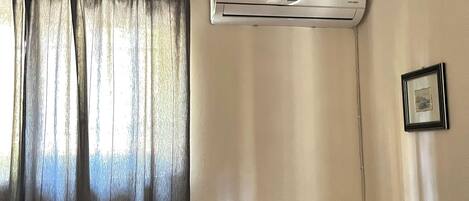 Una habitación de hotel con cama, unidad de aire acondicionado, mesita de noche con lámpara y un cuadro en la pared.