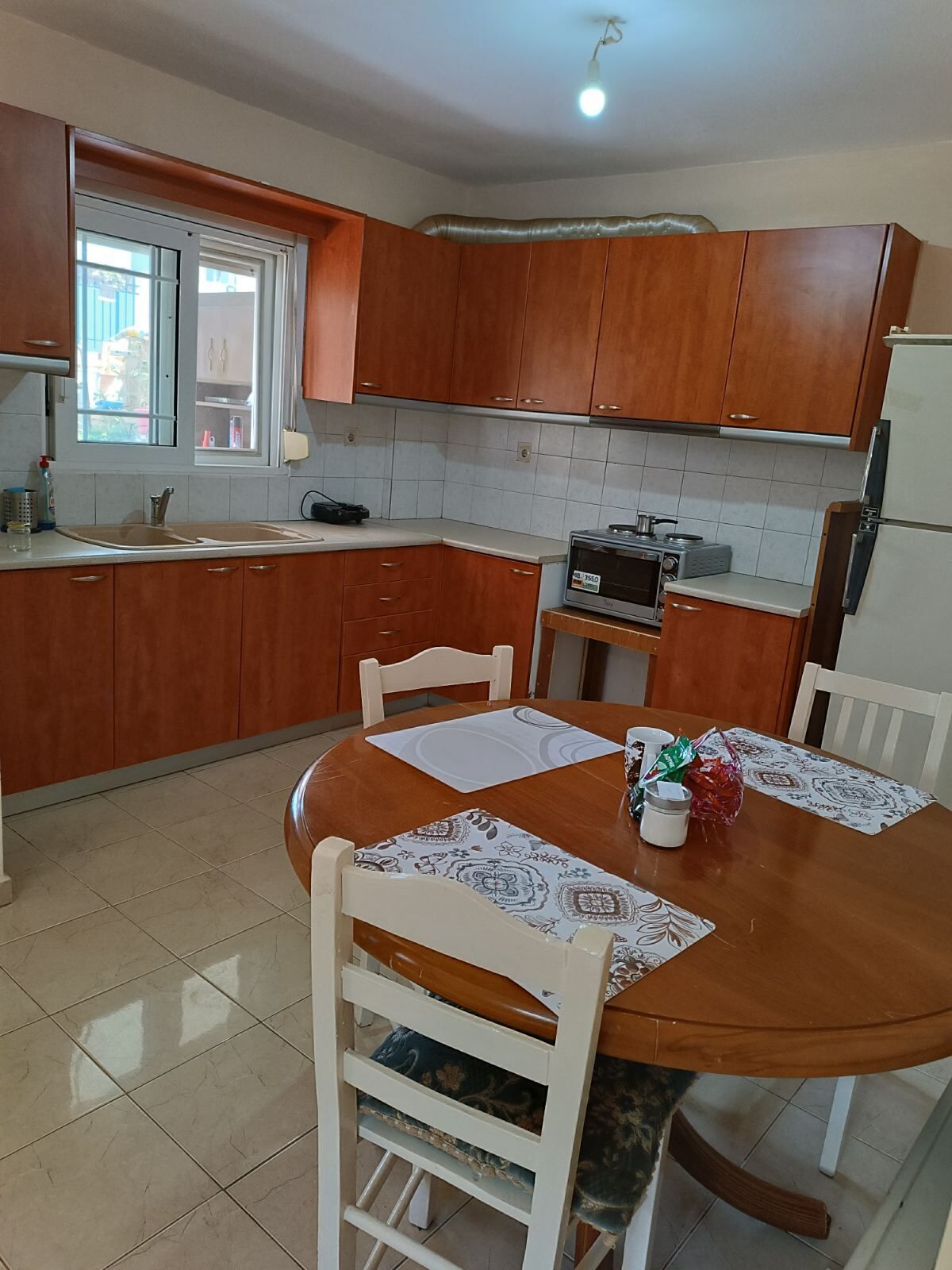 A kitchen with wooden cabinets, a microwave, a refrigerator, a dining table with chairs, and a window with a view of the outside.