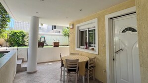 Uma área de varanda coberta com mesa e cadeiras, uma porta branca e uma janela com vista para o exterior.