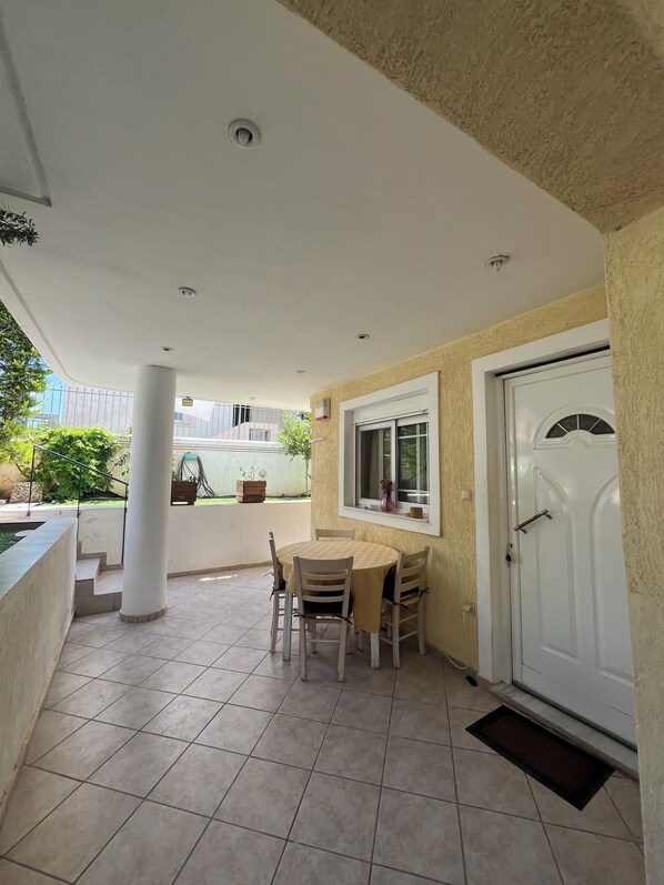 A covered patio area with a table and chairs, a white door, and a window with a view of the outside. - Stavros Apartments (Spata-Artemida)