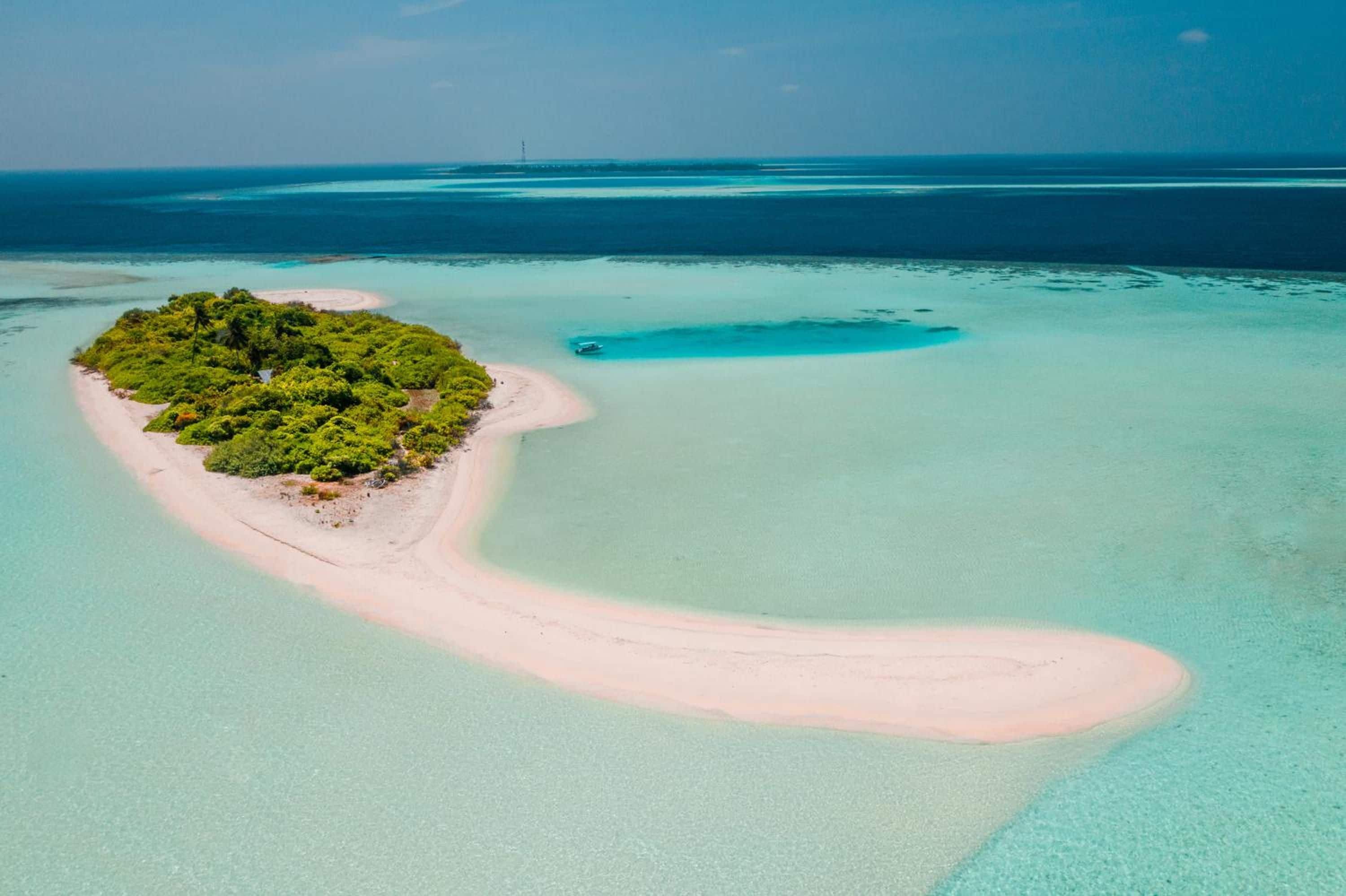 An aerial view of a tropical island with a narrow sandy beach, dense green vegetation, and clear turquoise waters.
