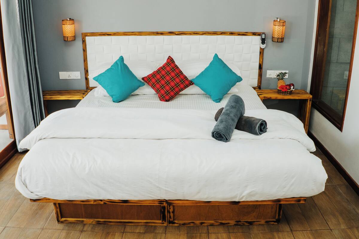 Una cama grande con cabecera blanca, almohadas turquesa y rojas, y toallas enrolladas a un lado.