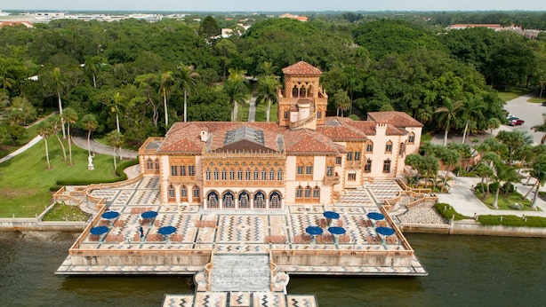 Un edificio imponente e decorato, con una torre centrale, circondato da un elemento d'acqua e palme.