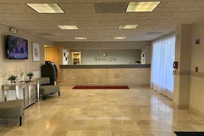 A reception area with a tiled floor, a reception desk, a television, and a wall-mounted sign that reads "RAMADA".