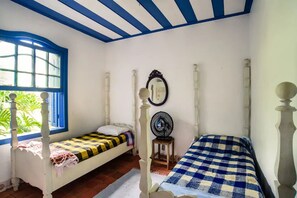 A room with two single beds, a window with blue shutters, a mirror, and a small table with a fan. - Ilha Bela a Beira Mar (Ilhabela)