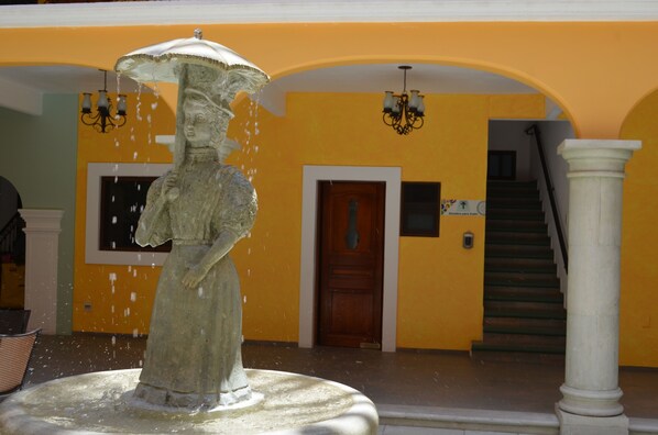 A fountain with a statue of a woman holding an umbrella, set against a yellow wall with a staircase and a door in the background. - Hotel Casa Franco (San Pedro y San Pablo Teposcolula)