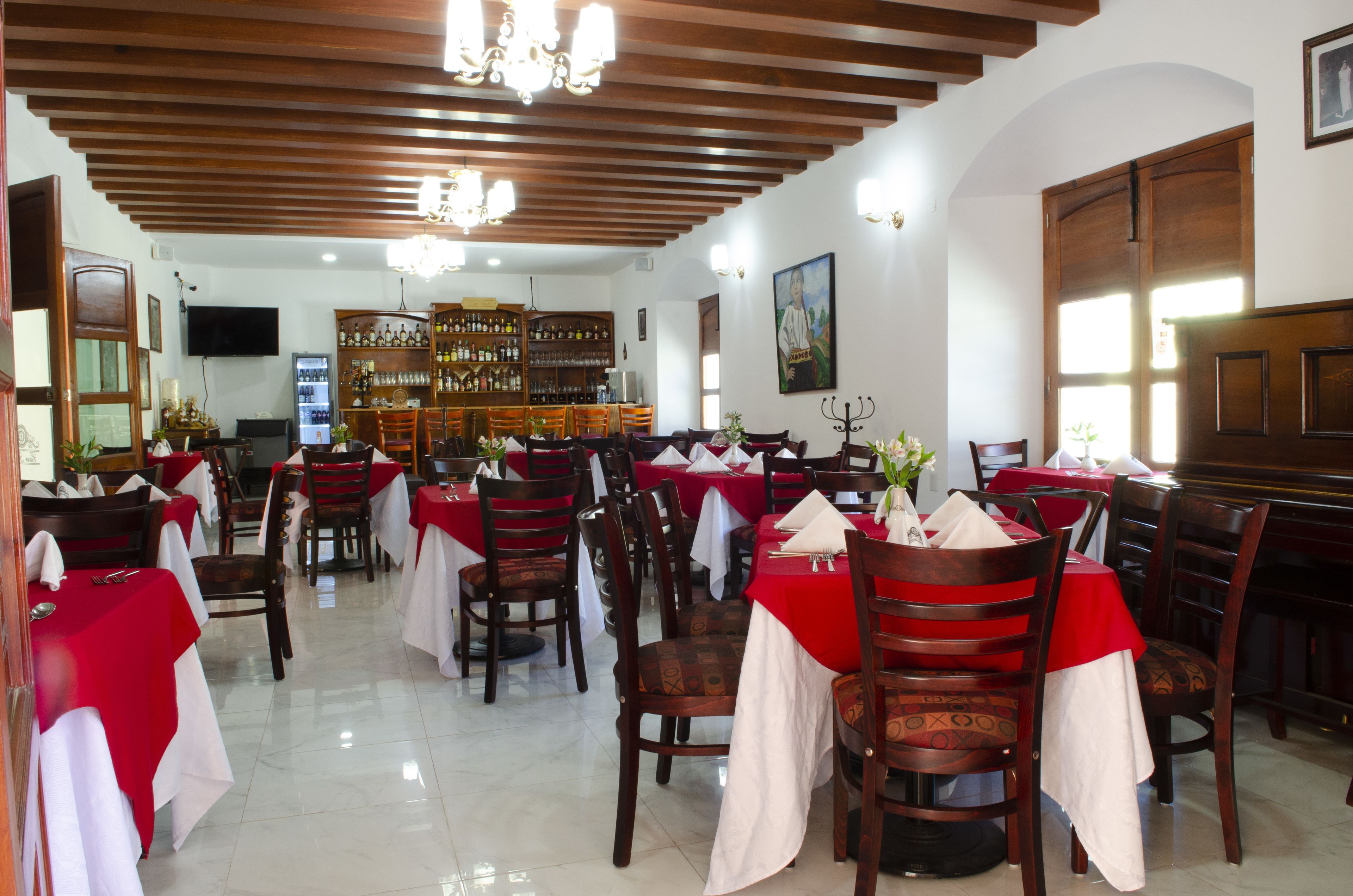 A restaurant with wooden ceiling beams, red tablecloths, and a bar area with shelves of bottles.