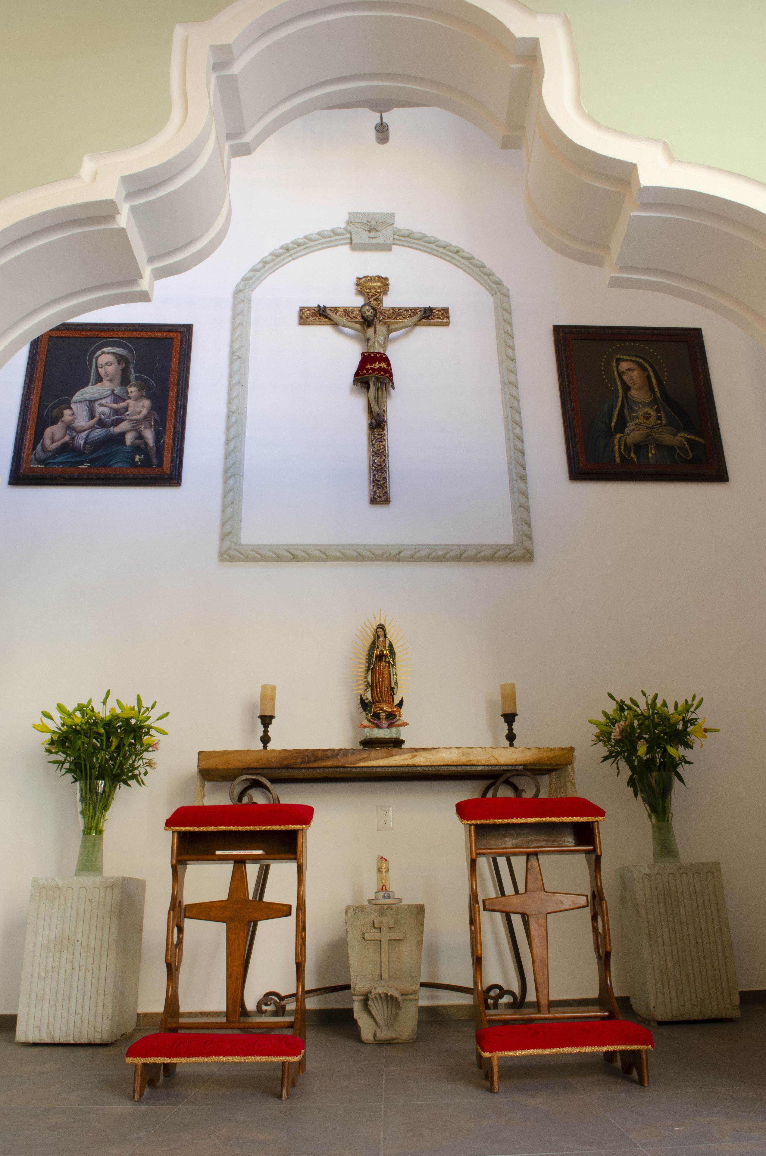 Innenansicht einer Kirche mit Kruzifix, zwei Holzbänken und gerahmten religiösen Bildern.