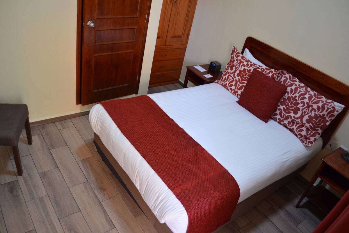 A neatly made bed with a red and white bedspread, a wooden headboard, and a nightstand with a lamp.