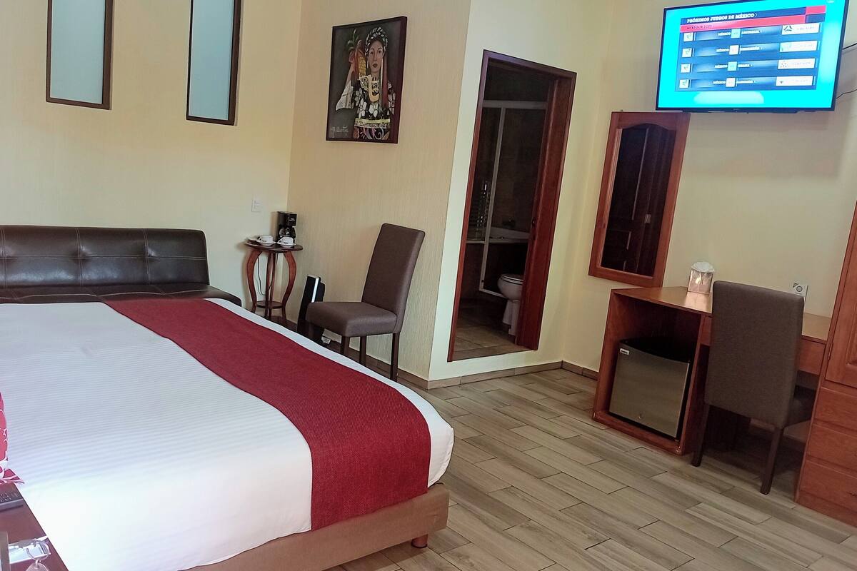 A hotel room with a large bed, a desk, a chair, a TV, and a wardrobe.