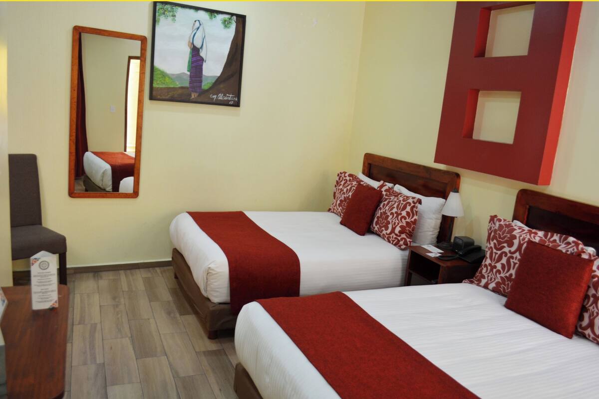 A hotel room with two beds, a mirror, a nightstand, a desk, and a painting on the wall.