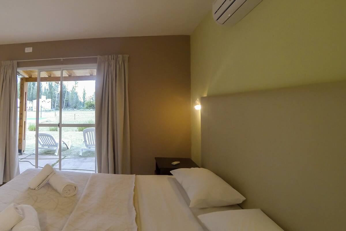 Habitación de hotel con una cama grande, una mesita de noche y vistas al exterior a través de un ventanal corredero.