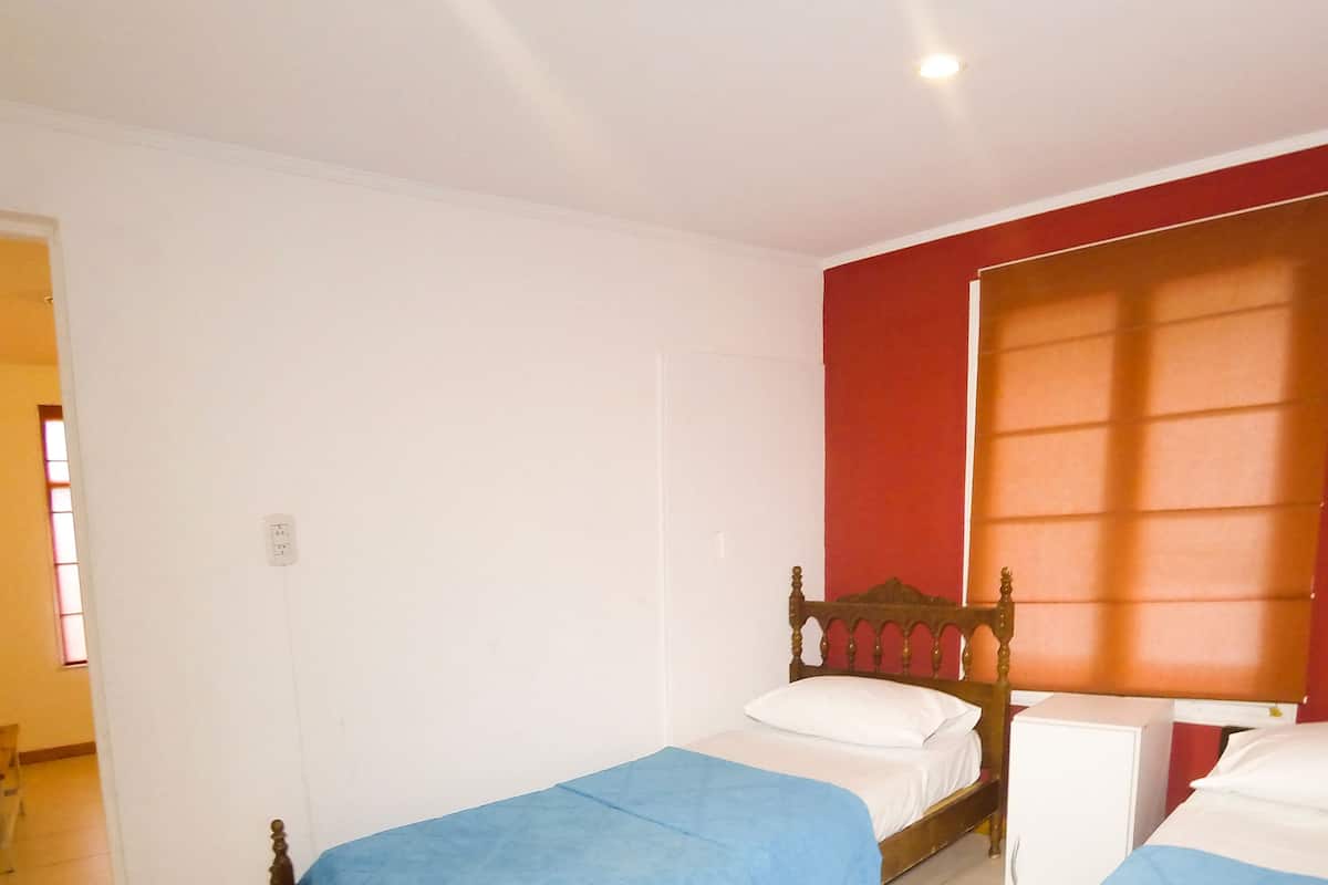 Habitación con dos camas individuales, cabecero de madera, ventana con persianas y puerta que da a otra habitación.