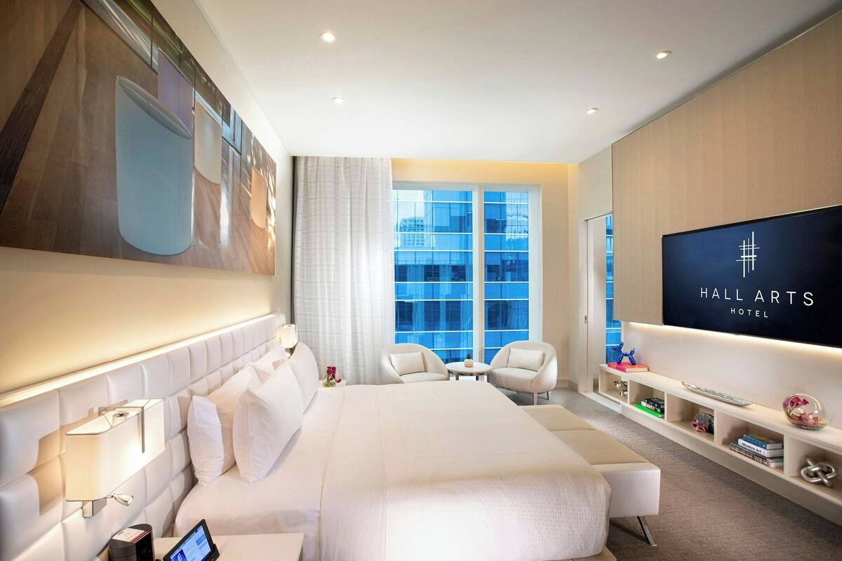 A modern hotel room with a large bed, a TV, a desk, and a seating area with chairs.