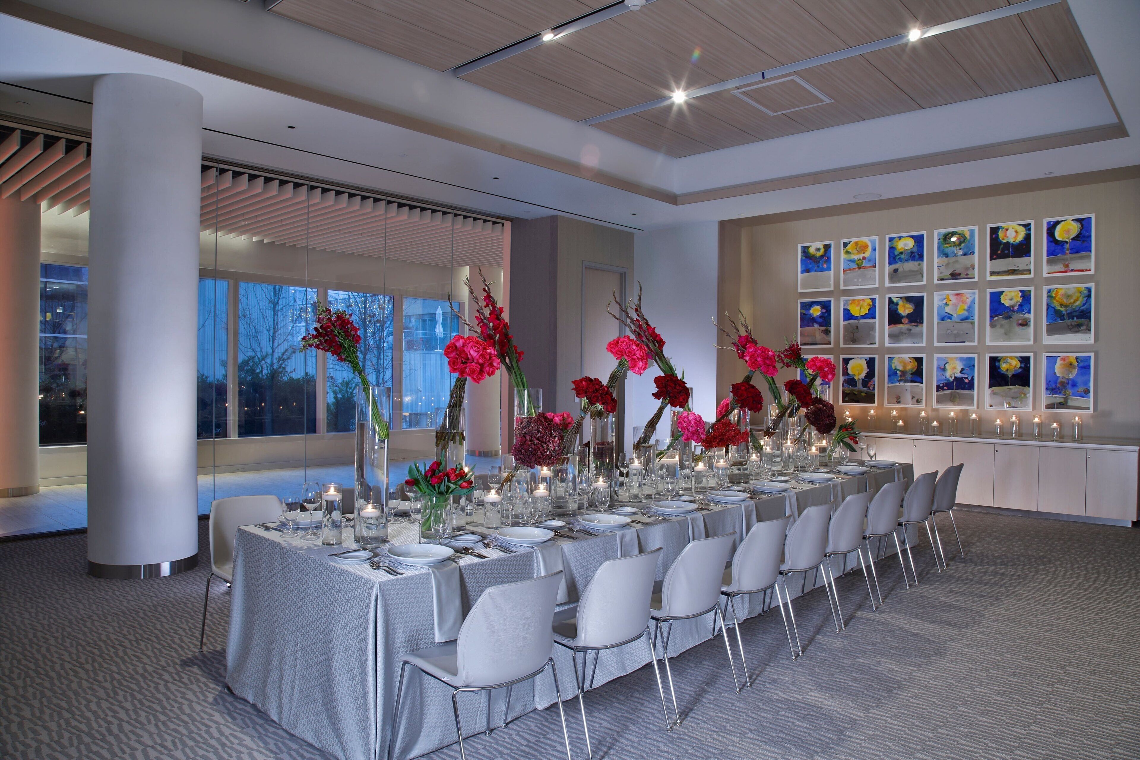 Uma longa mesa de jantar posta para um evento formal, com cadeiras brancas e centros de mesa florais.