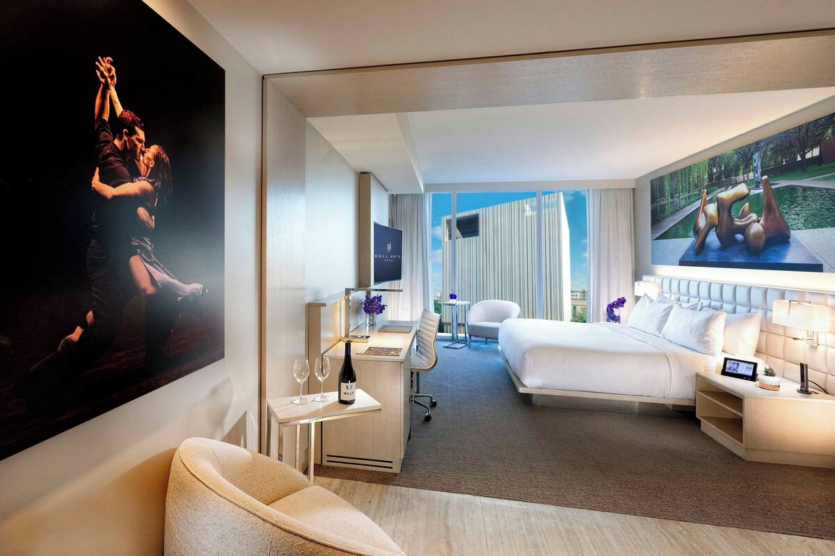 A modern hotel room with a large bed, a desk with a chair, a seating area, and artwork on the walls.