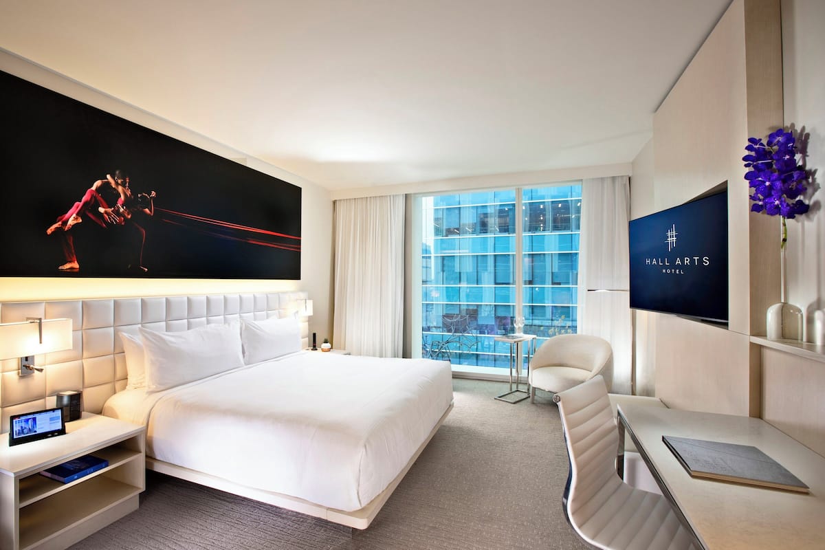 A modern hotel room with a large bed, a desk, a chair, a TV, and a window with a city view.