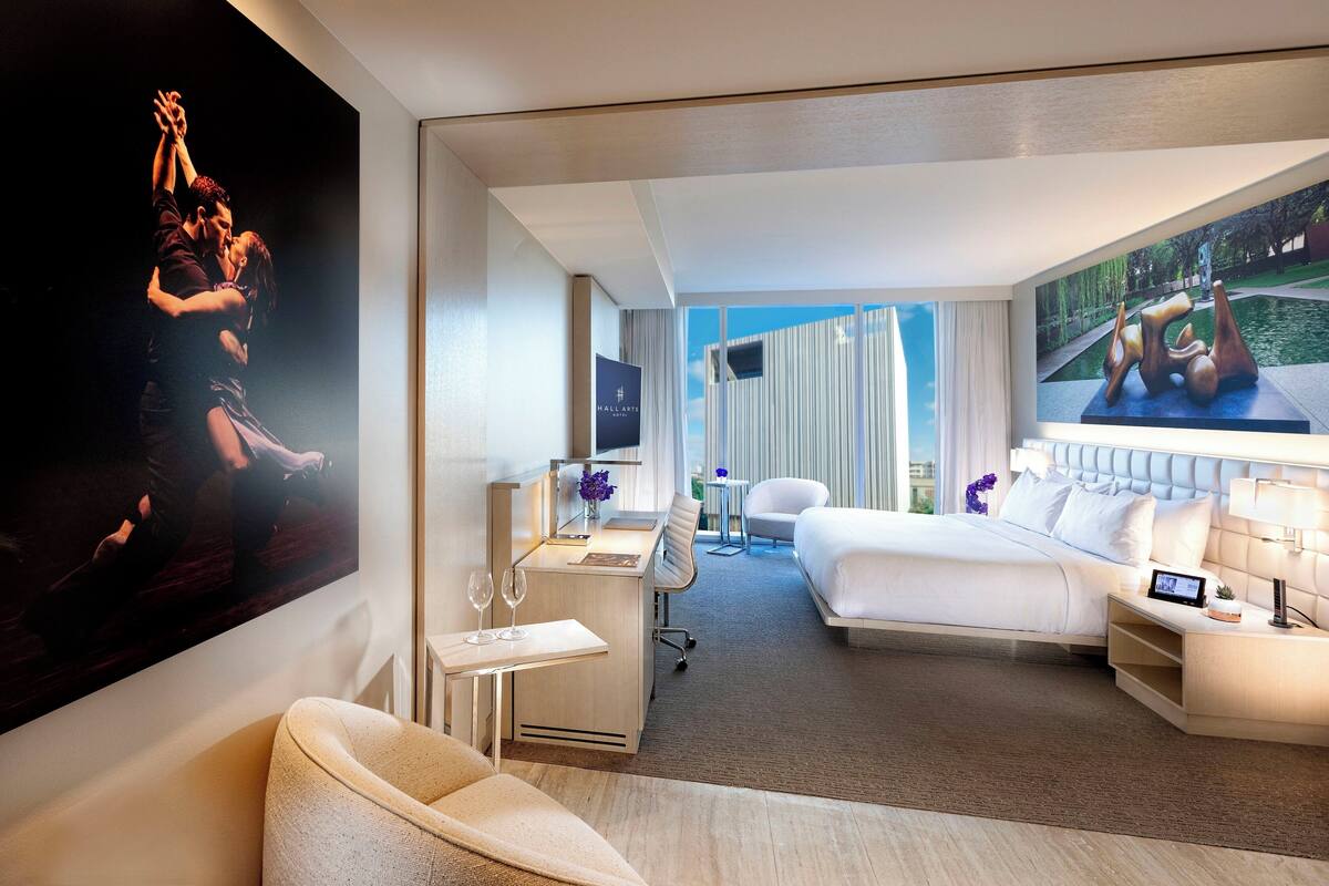 A hotel room with a bed, a desk, a chair, a TV, and a large painting on the wall.