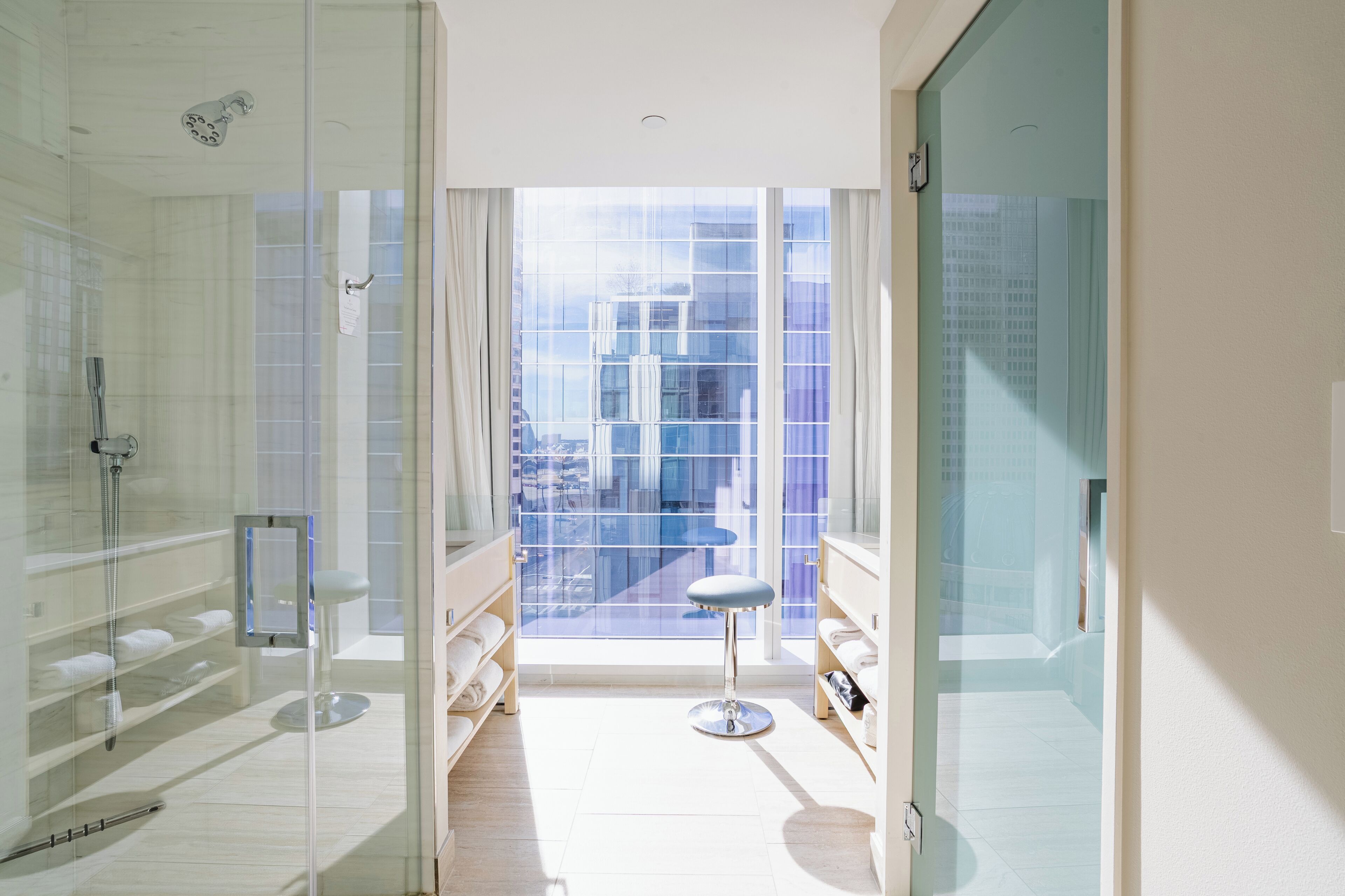 A modern bathroom with a glass shower enclosure, a vanity with a mirror, and a view of a cityscape through large windows.