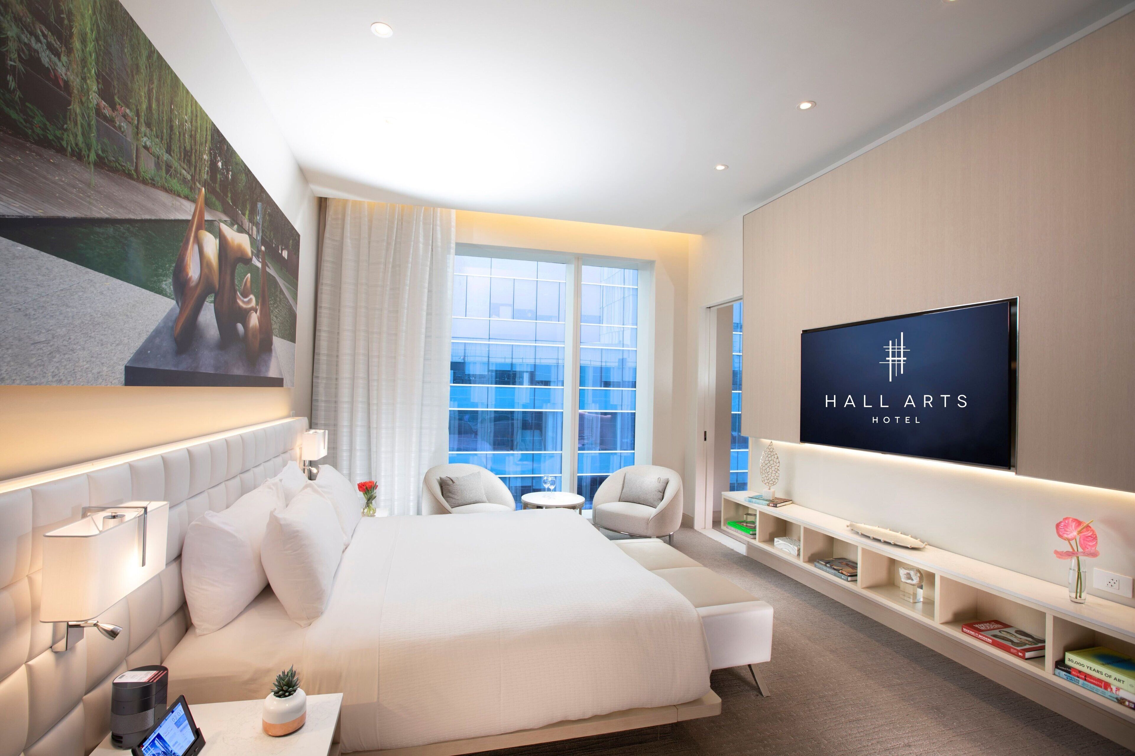 A modern hotel room with a large bed, a flat-screen TV, a seating area, and a view of the outdoors.