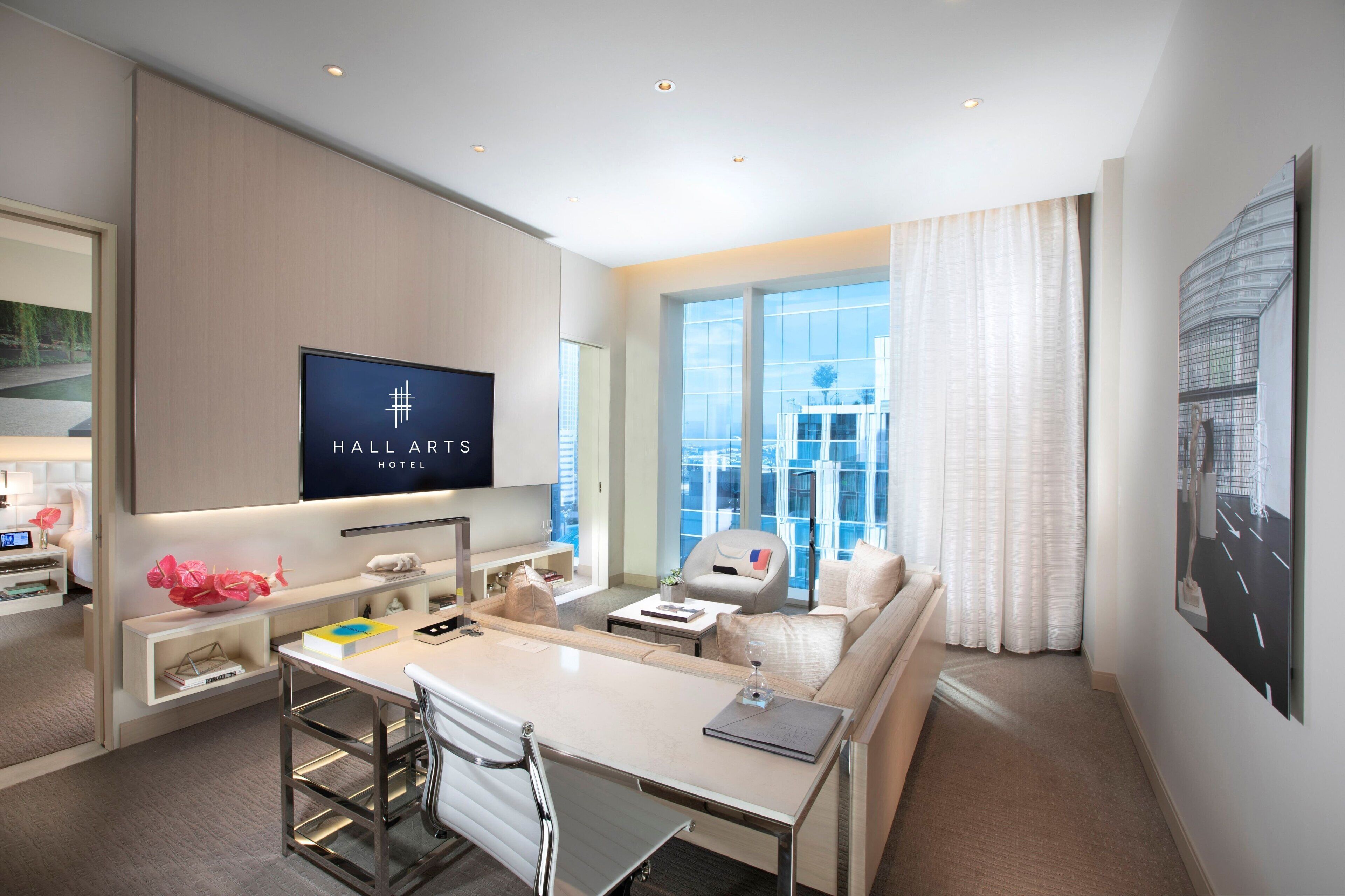 Una habitación de hotel moderna con comedor, sofá, televisión y un amplio ventanal con vista a la ciudad.