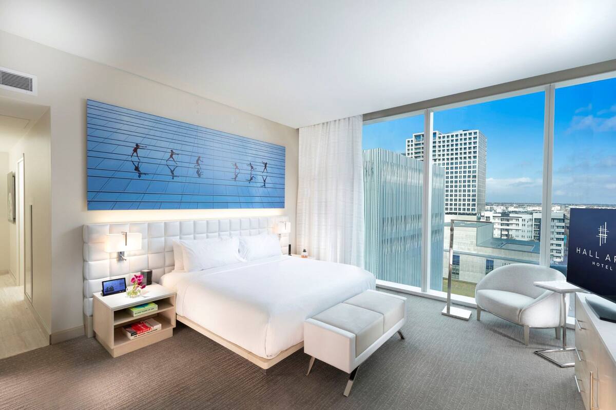 A modern hotel room with a large bed, a bedside table with a lamp, a small table with a chair, and a large window offering a city view.