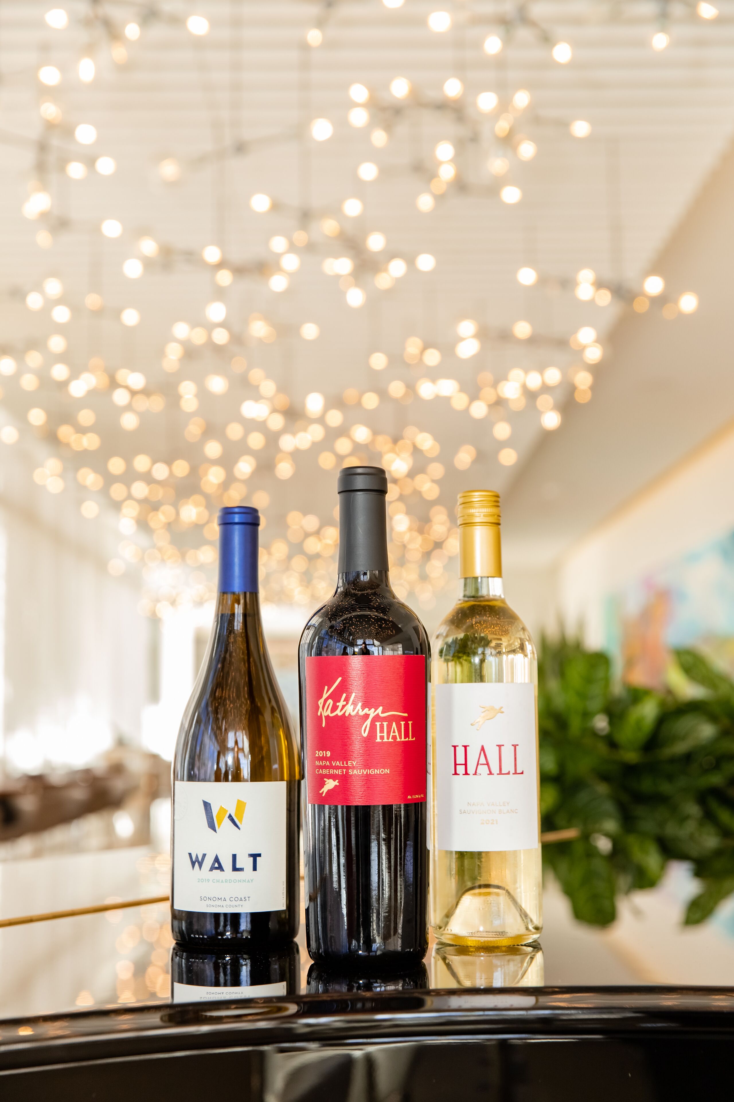 Three wine bottles on a table with a ceiling of string lights.